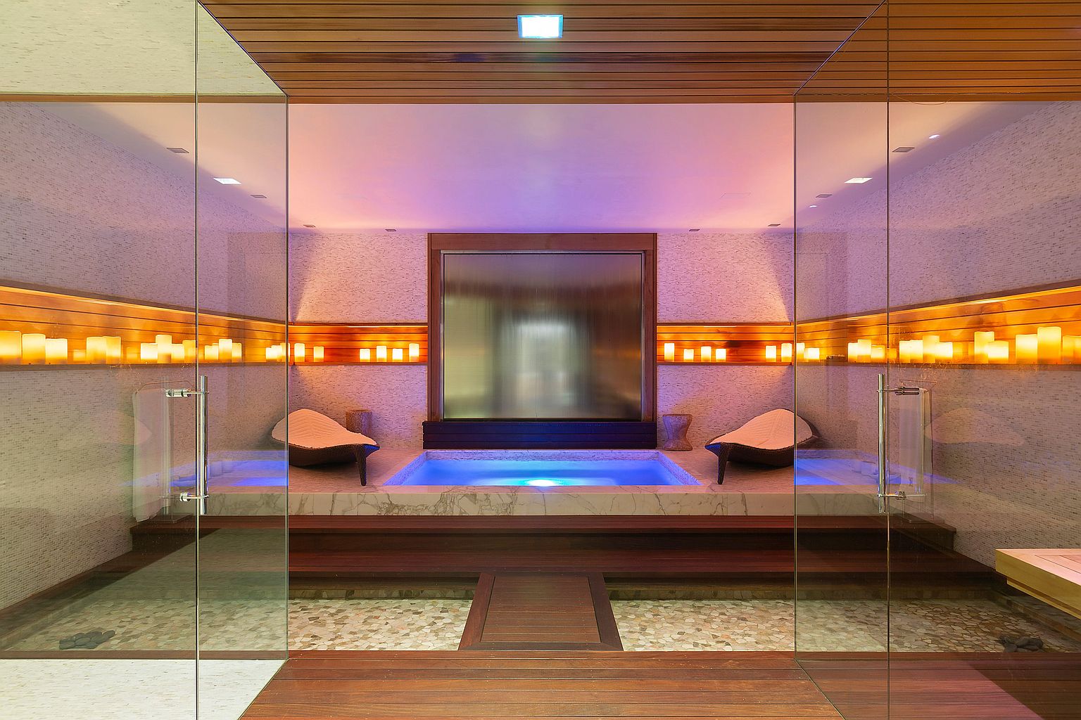 This luxurious primary bathroom features a modern spa area with a hot tub illuminated by blue light, a waterfall feature, and ambient candle lighting. The space is enclosed by glass walls, showcasing the wood and stone finishes, creating a serene and upscale atmosphere. The perspective is from the entrance of the spa.