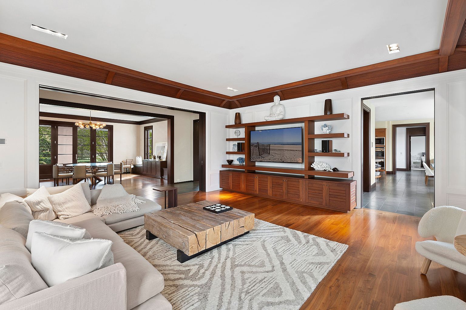 This is an interior shot of a luxurious living room featuring hardwood floors, a large sectional sofa with plush pillows, and a rustic wooden coffee table. A built-in entertainment center with a large television and decorative shelving serves as a focal point, while open doorways lead to adjacent rooms, enhancing the sense of spaciousness and flow. The room is well-lit and exudes a comfortable yet sophisticated ambiance.
