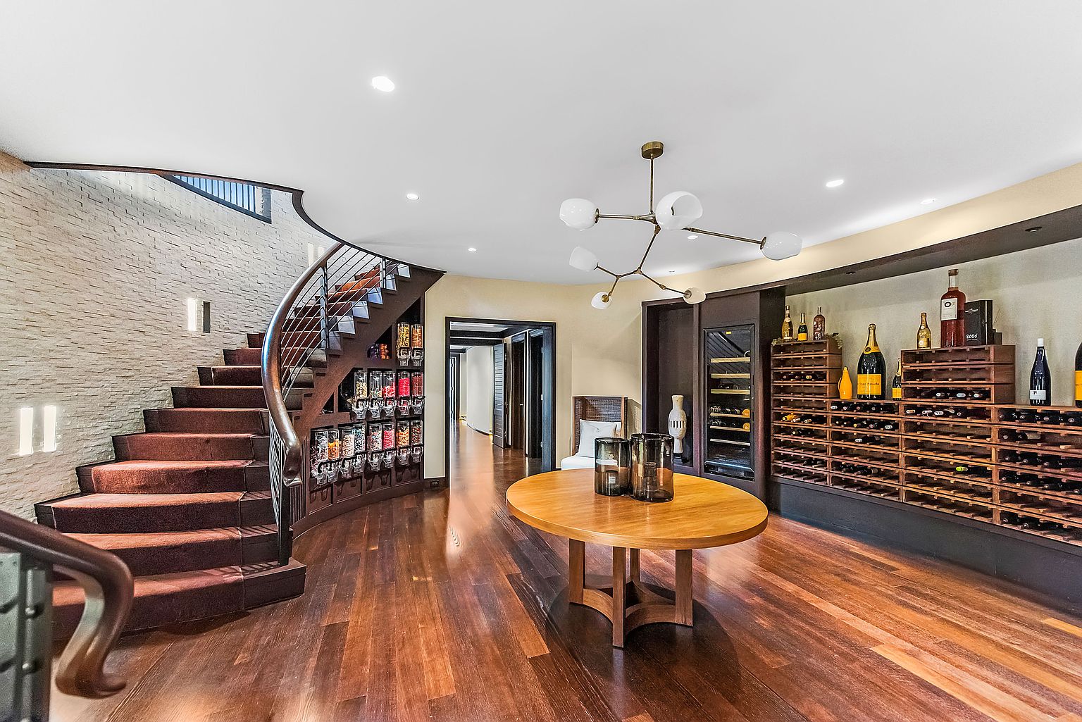 This interior shot showcases a sophisticated wine cellar with a curved staircase and a custom wine rack display. A round wooden table sits in the center of the room, illuminated by a modern chandelier. The hardwood flooring and brick accent wall add to the room's luxurious and inviting atmosphere.