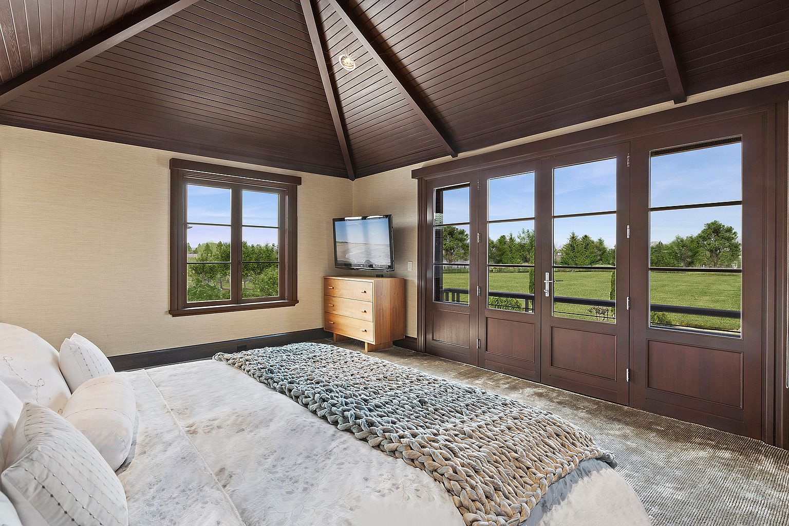 This is a primary bedroom featuring a bed with white linens and a textured throw blanket. The room has a vaulted ceiling with dark wood beams, a window, and a set of French doors leading to an outdoor view. A wooden dresser with a television on top sits against the wall, and the overall style is luxurious and comfortable.