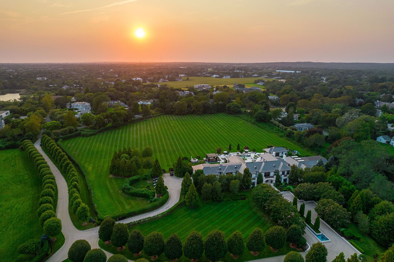 This aerial view showcases a sprawling estate at sunset, featuring a meticulously manicured lawn with striped patterns, a grand residence with a gray roof, and a long, tree-lined driveway. The property is surrounded by lush greenery and other homes, creating a sense of privacy and exclusivity. The warm hues of the setting sun add a touch of luxury and tranquility to the scene.