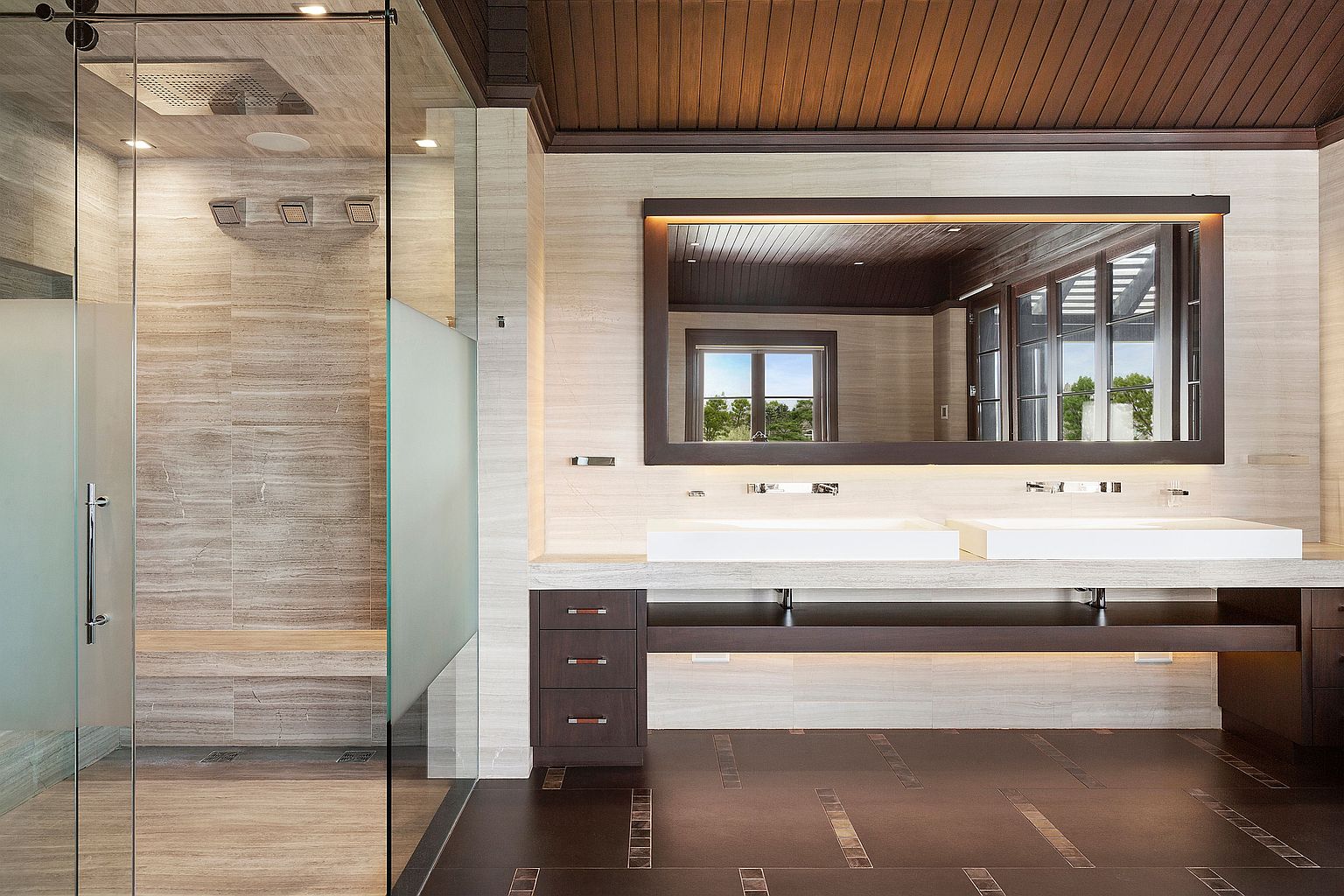 This is a luxurious primary bathroom featuring a spacious walk-in shower with multiple shower heads and a built-in bench. The double vanity has modern, rectangular sinks and a large mirror with integrated lighting. The flooring is a combination of dark and light tiles, adding visual interest to the space.