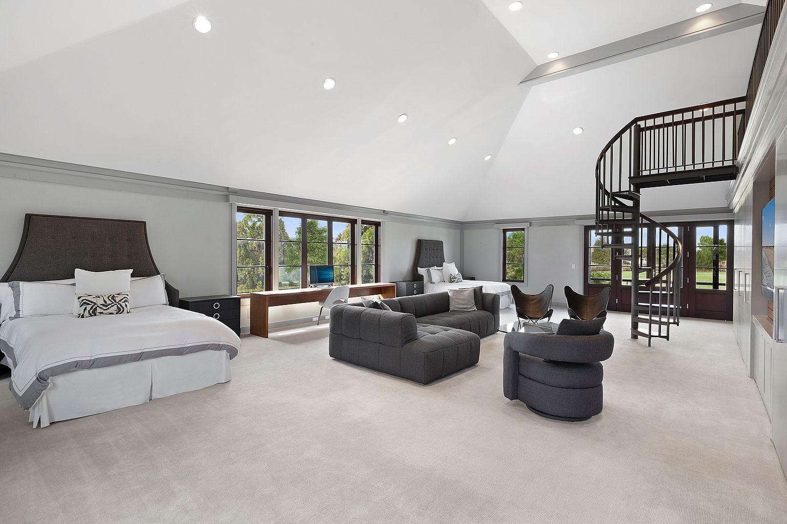 This is a spacious primary bedroom featuring two beds, a spiral staircase, and a seating area with a gray sofa and chairs. The room has large windows that provide natural light and views of the outdoors. The overall style is modern and luxurious, with a neutral color palette and high ceilings.