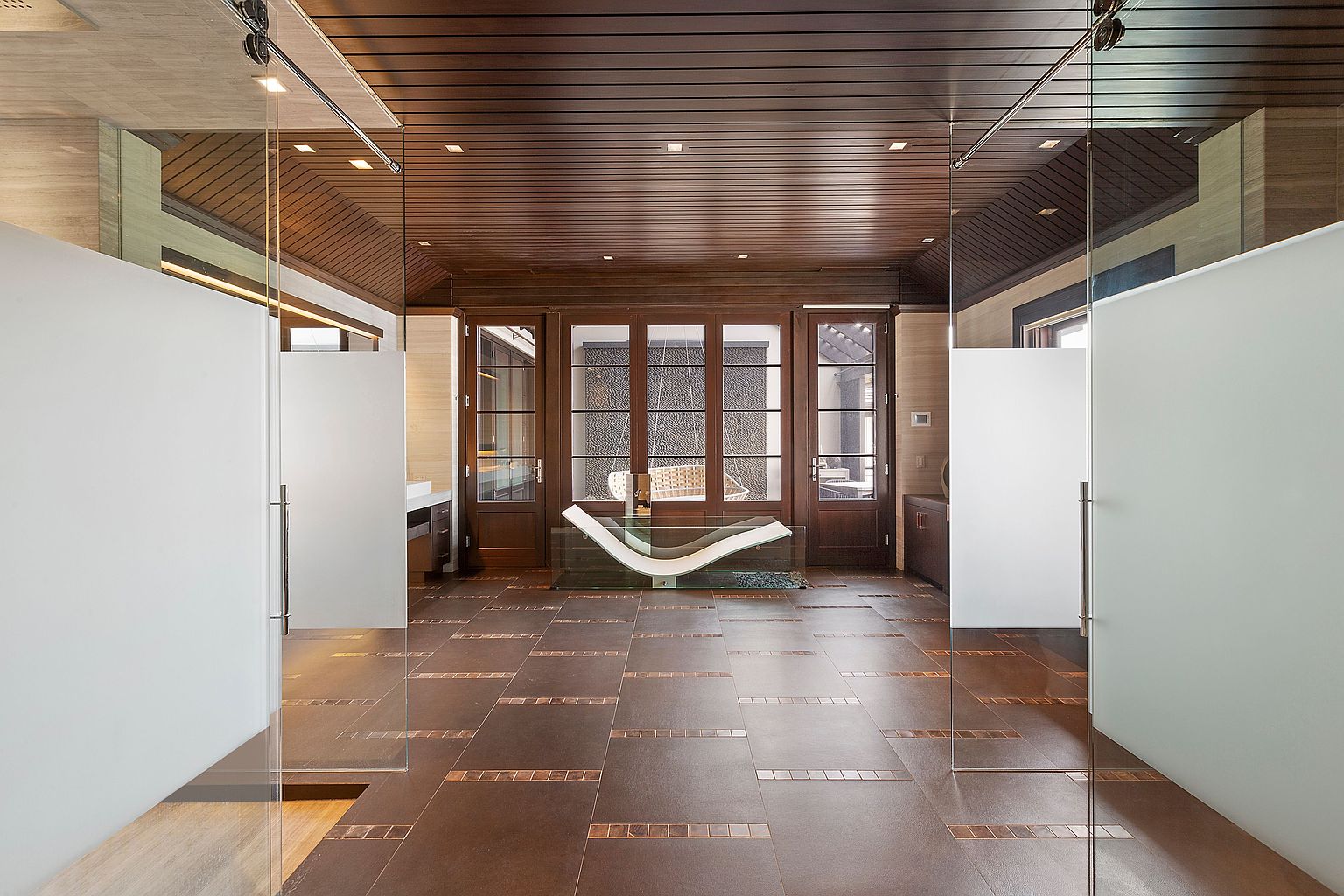 This is a luxurious primary bathroom featuring a modern design with dark wood ceilings and tiled floors. The room is divided by frosted glass panels, creating separate spaces while maintaining an open feel. A unique, curved bench sits in front of a set of doors leading to an outdoor area, adding a touch of elegance and relaxation.
