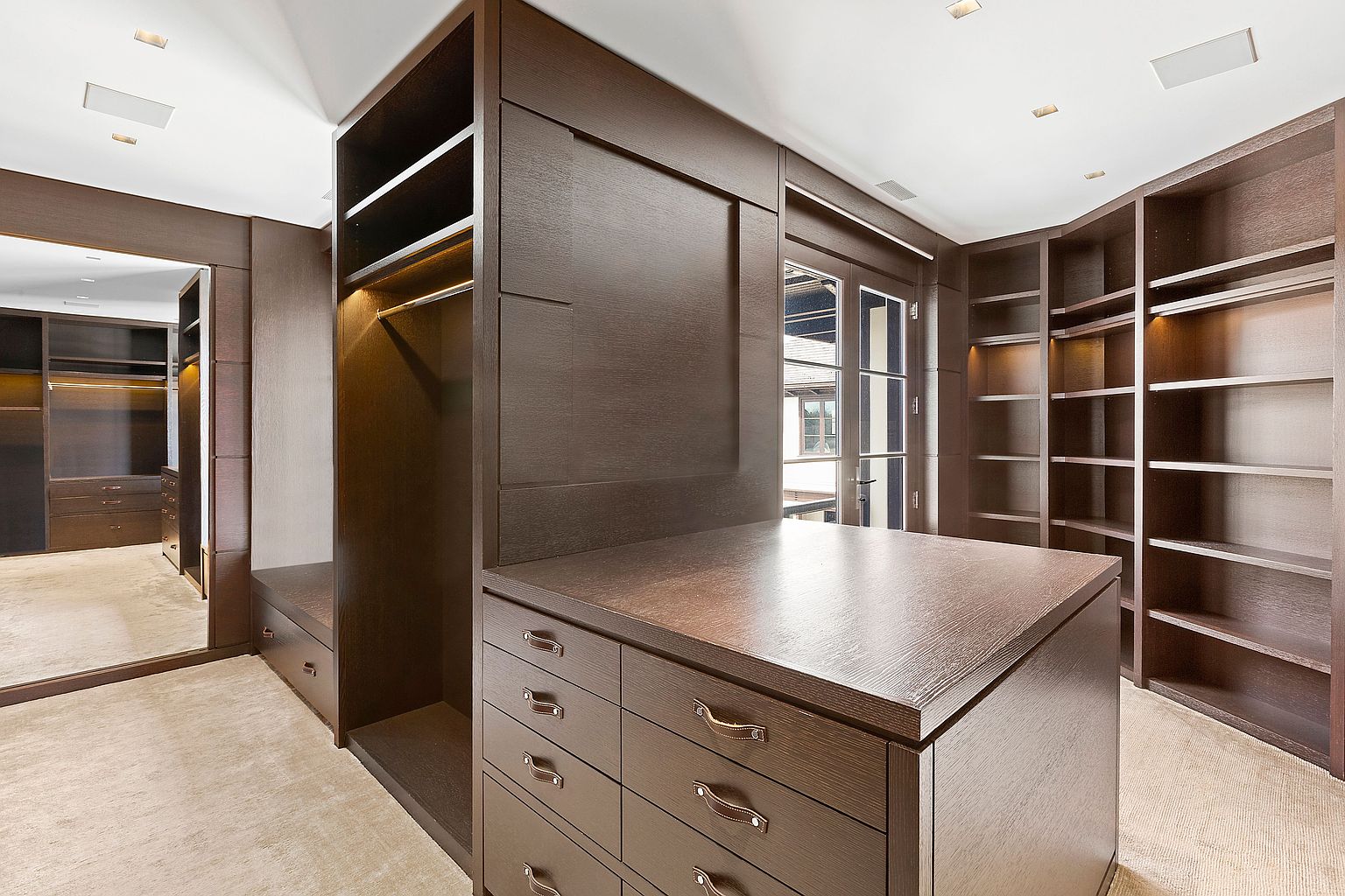 This is a well-organized walk-in closet featuring dark wood cabinetry and shelving. An island with drawers sits in the center, providing additional storage and workspace. A full-length mirror is visible on the left, and natural light enters through a glass door, creating a luxurious and functional space.