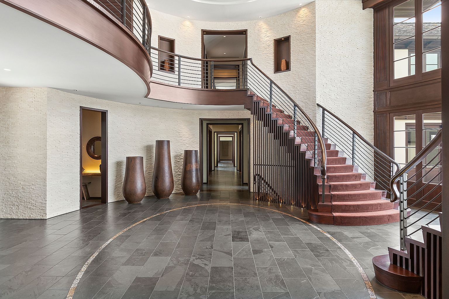 This grand foyer features a curved staircase with a wrought iron railing and carpeted steps, complemented by a second-story balcony overlooking the entrance. The flooring is a dark tile with a circular accent, and the walls are textured in a light color. Three large, decorative vases add a touch of elegance to the space, creating a luxurious and inviting atmosphere.