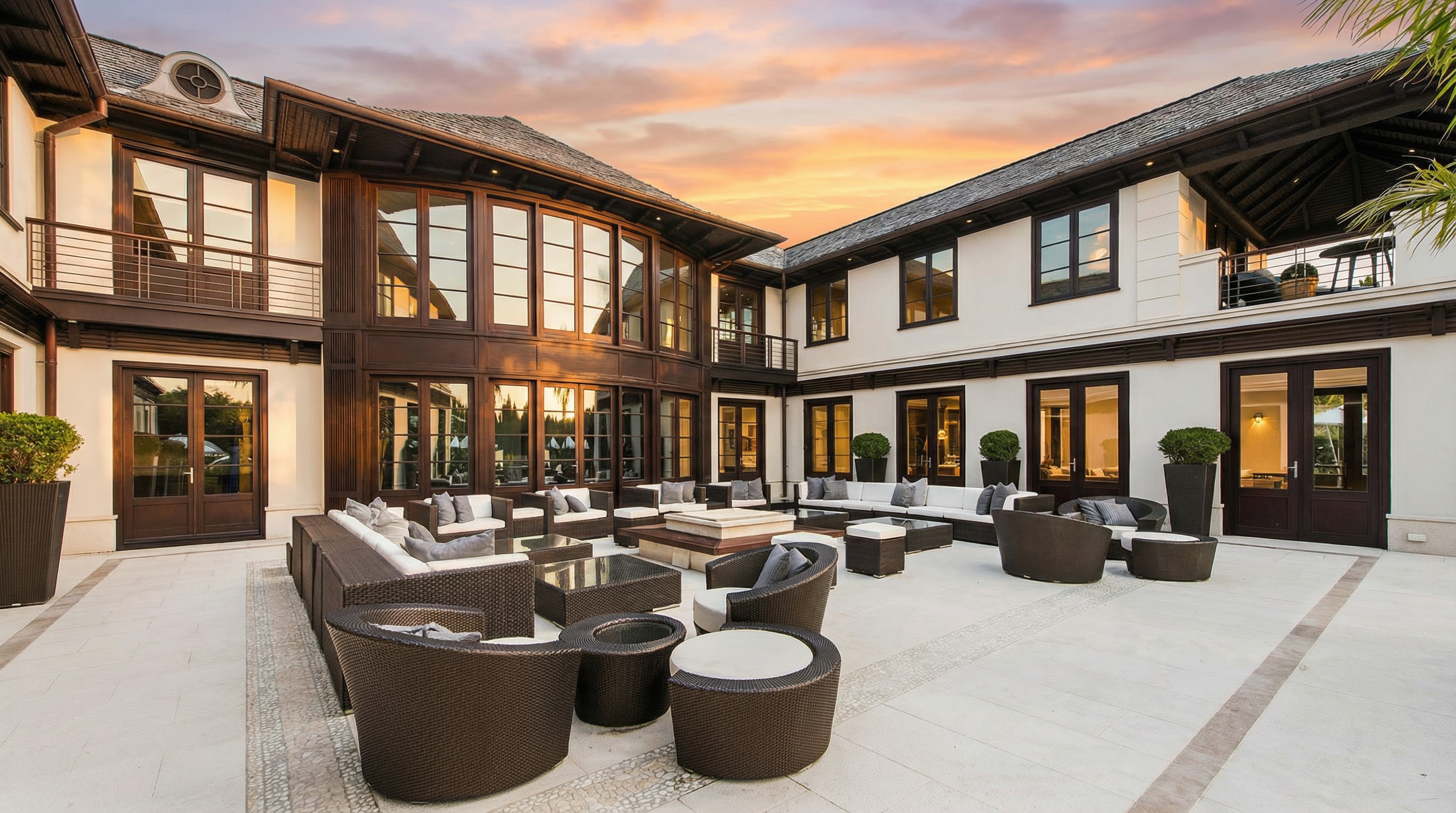 This is an exterior shot of a luxurious patio area, featuring elegant outdoor furniture arranged around a central fire pit. The patio is surrounded by a grand house with large windows and doors, suggesting a seamless indoor-outdoor living experience. The overall impression is one of sophistication and comfort, perfect for entertaining or relaxing.
