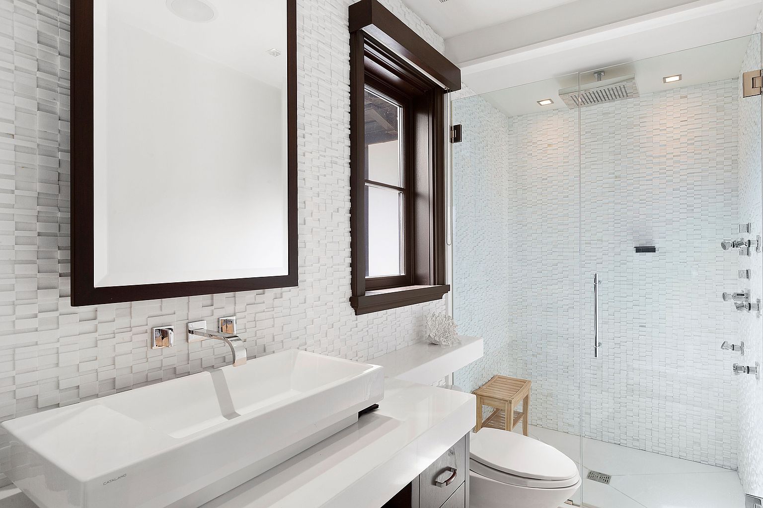 This is a modern primary bathroom featuring a sleek, rectangular white sink atop a white countertop with dark wood accents on the mirror and window frame. The walls are adorned with textured white tiles, and a glass-enclosed shower with mosaic tile walls is visible in the background. The overall impression is clean, contemporary, and luxurious.