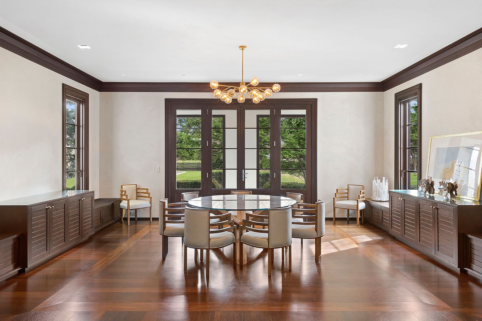 This is a spacious dining room featuring a round glass-top table surrounded by upholstered chairs. The room is well-lit by a modern chandelier and natural light from large windows and doors that offer a view of the exterior landscape. Dark wood trim and built-in cabinets add a touch of elegance and sophistication to the space.