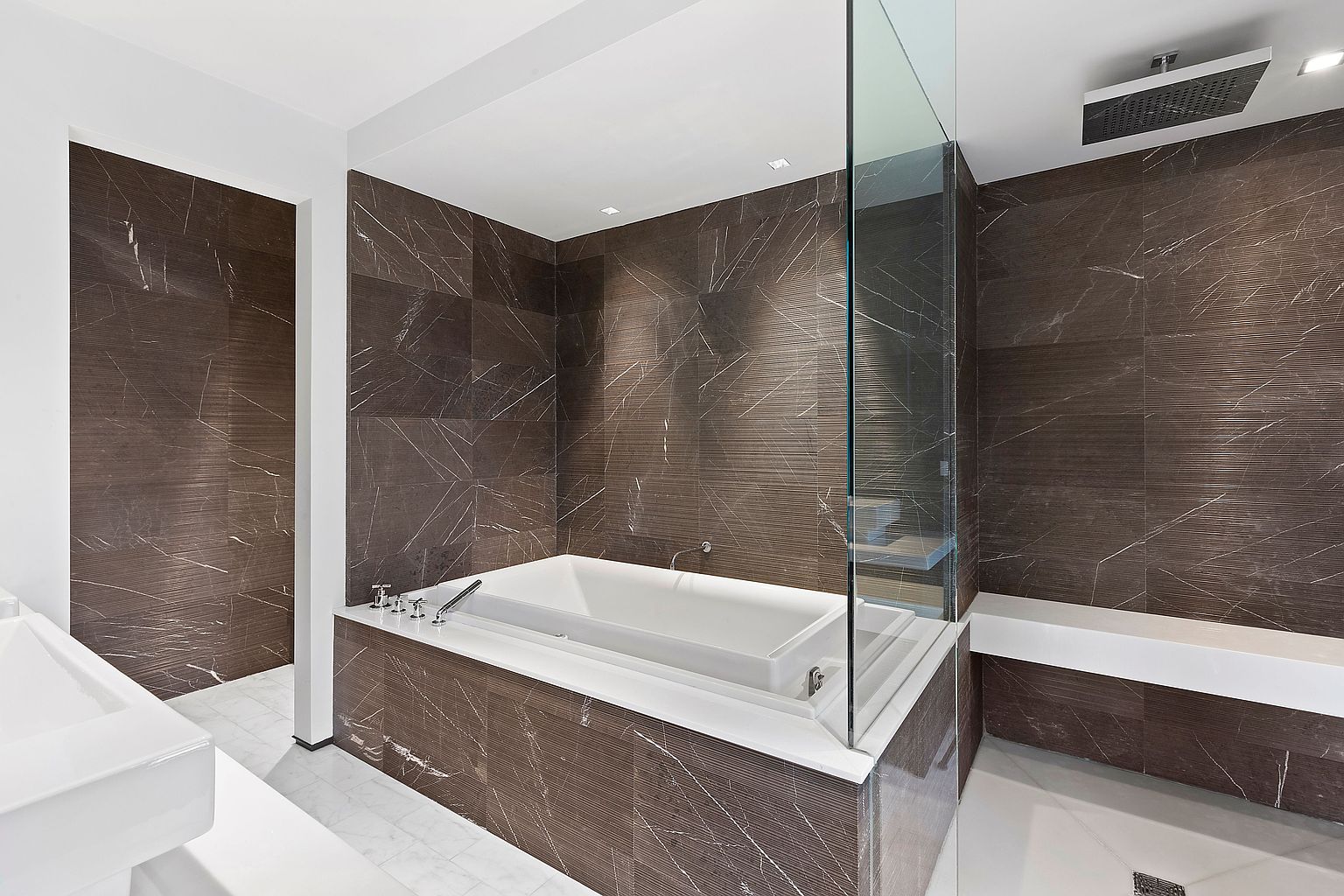 This is a luxurious primary bathroom featuring dark brown marble-like tile walls and white marble flooring. A modern bathtub is encased in the same dark tile with a white countertop surround, and a glass partition separates the tub from the shower area. The space is well-lit with recessed lighting, creating a spa-like atmosphere.