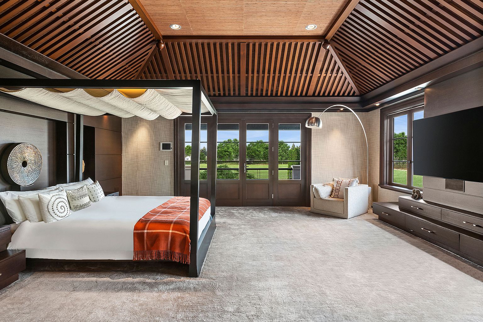 This is a luxurious primary bedroom featuring a four-poster bed with a draped canopy, complemented by an orange throw blanket. The room boasts a unique wooden ceiling design, a large window with a view of the outdoors, and a modern entertainment center with a large television. The overall aesthetic is elegant and serene, creating a relaxing retreat.