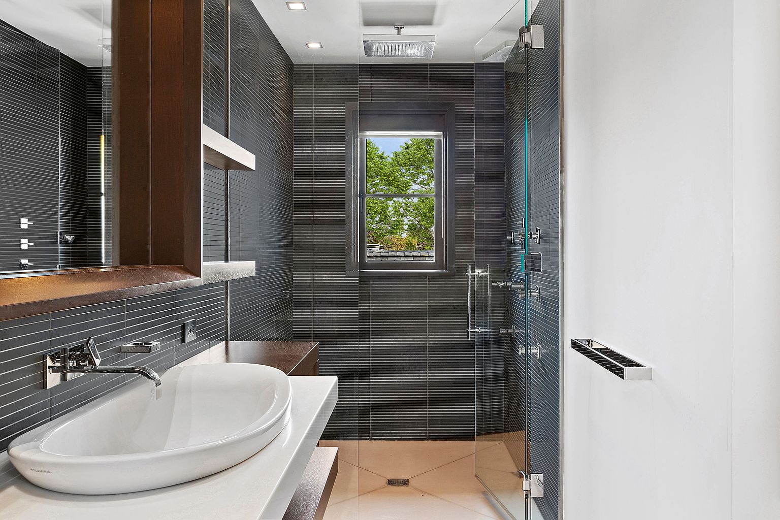 This is a modern bathroom featuring dark horizontal-striped tiles, a white vessel sink on a white countertop, and a glass-enclosed shower. A dark wood cabinet and a window providing natural light add to the contemporary design. The overall impression is sleek and sophisticated.