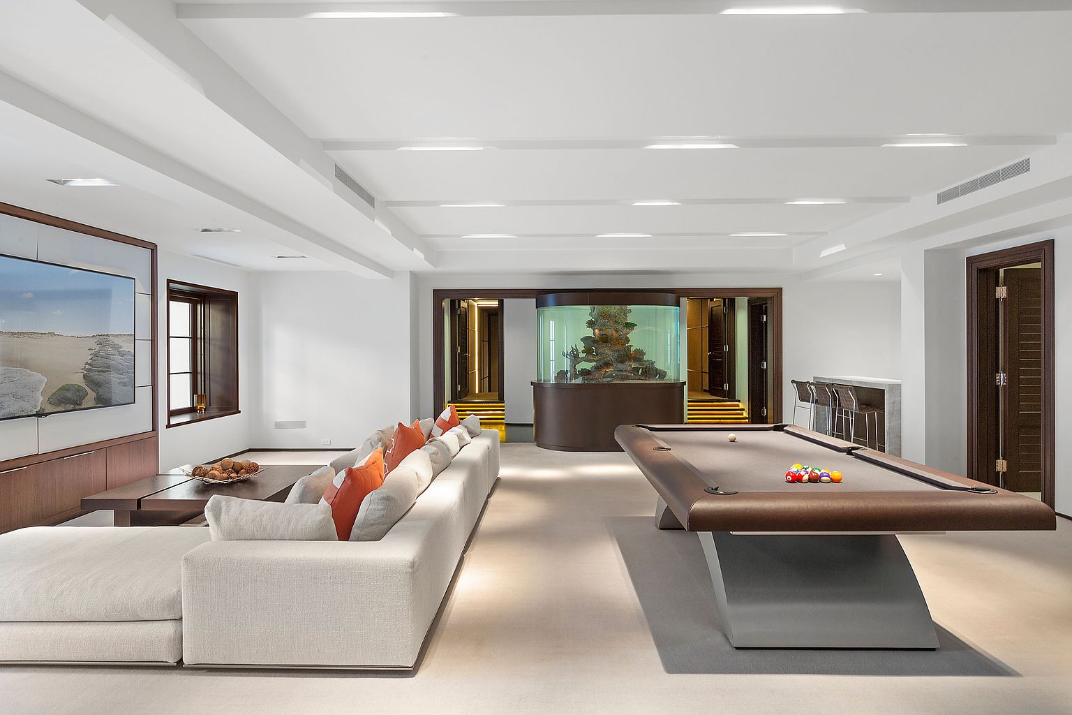 This is a spacious and luxurious living room featuring a large sectional sofa with orange accent pillows, a modern pool table, and a built-in aquarium. The room is well-lit with recessed lighting and natural light from a window, creating a bright and inviting atmosphere. The overall design is contemporary and elegant, perfect for entertaining and relaxation.