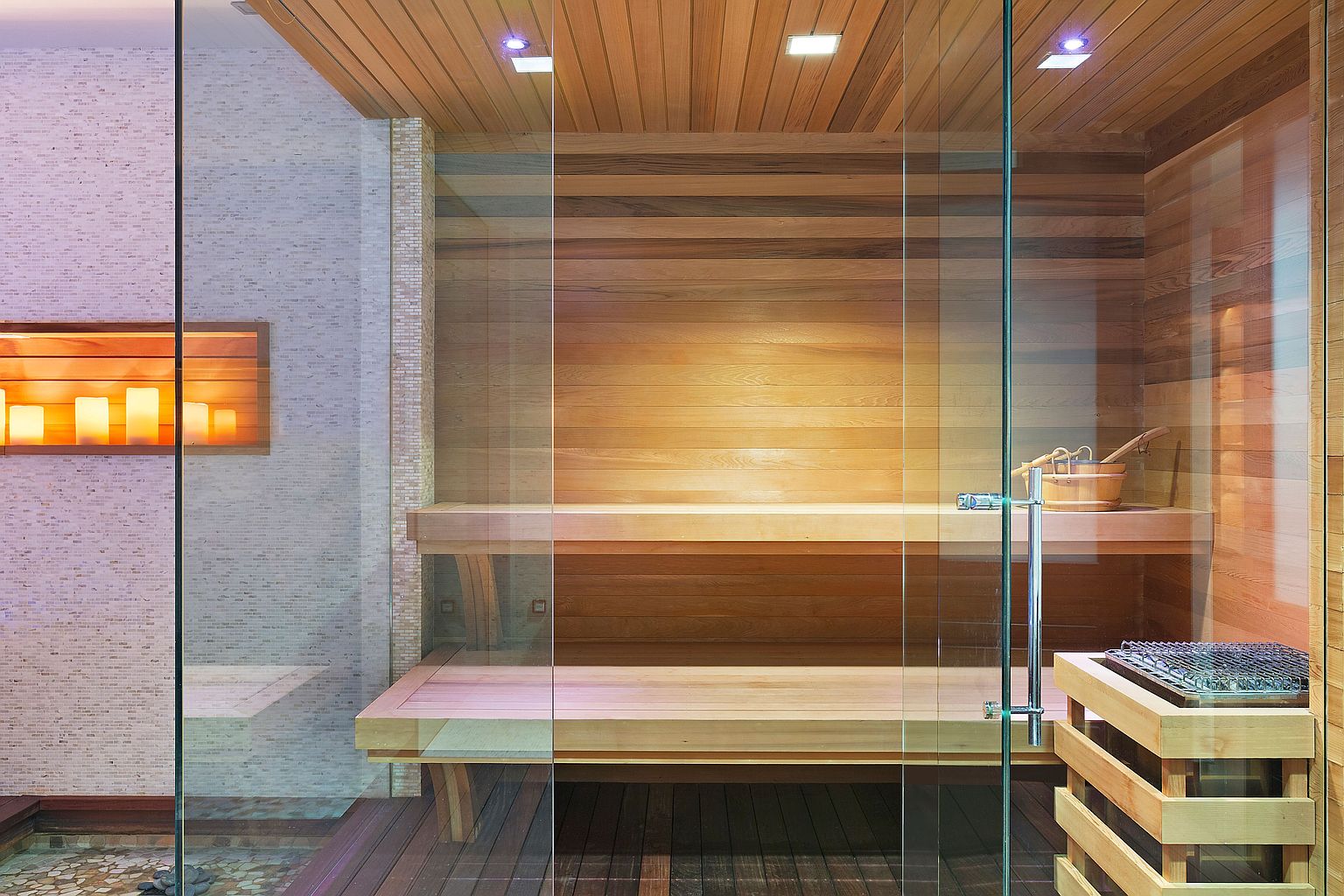 This image showcases a modern sauna, featuring wooden benches and walls, enclosed by glass panels. The sauna includes a wooden bucket and ladle, along with a heater, creating a spa-like atmosphere. The design emphasizes natural materials and clean lines, offering a luxurious and relaxing experience.