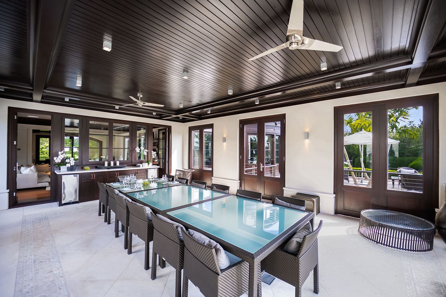 This image showcases a luxurious outdoor dining area on a covered patio. The space features a large glass-topped dining table surrounded by wicker chairs, a dark wood ceiling with recessed lighting and ceiling fans, and elegant dark wood framed doors leading to the interior and exterior. The overall impression is one of sophisticated outdoor living and entertaining.