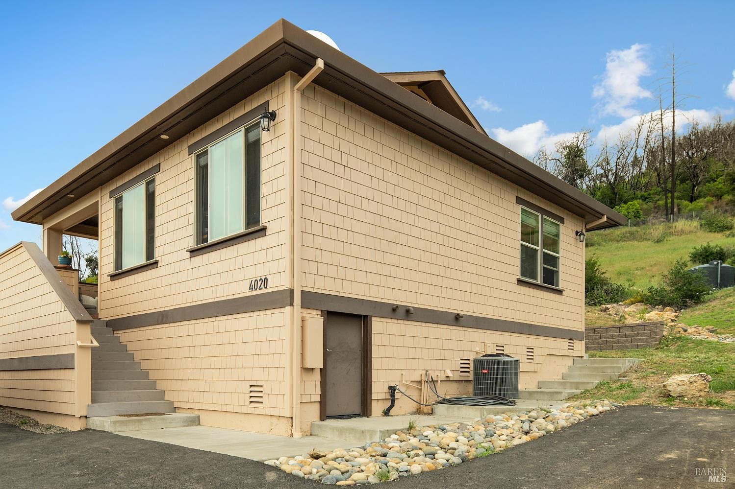 This is a side view of a two-story house with beige shingle siding and brown trim. The house features multiple windows, a ground-level entrance, and an external air conditioning unit. Landscaping includes decorative rocks and a paved driveway, with a hillside visible in the background.