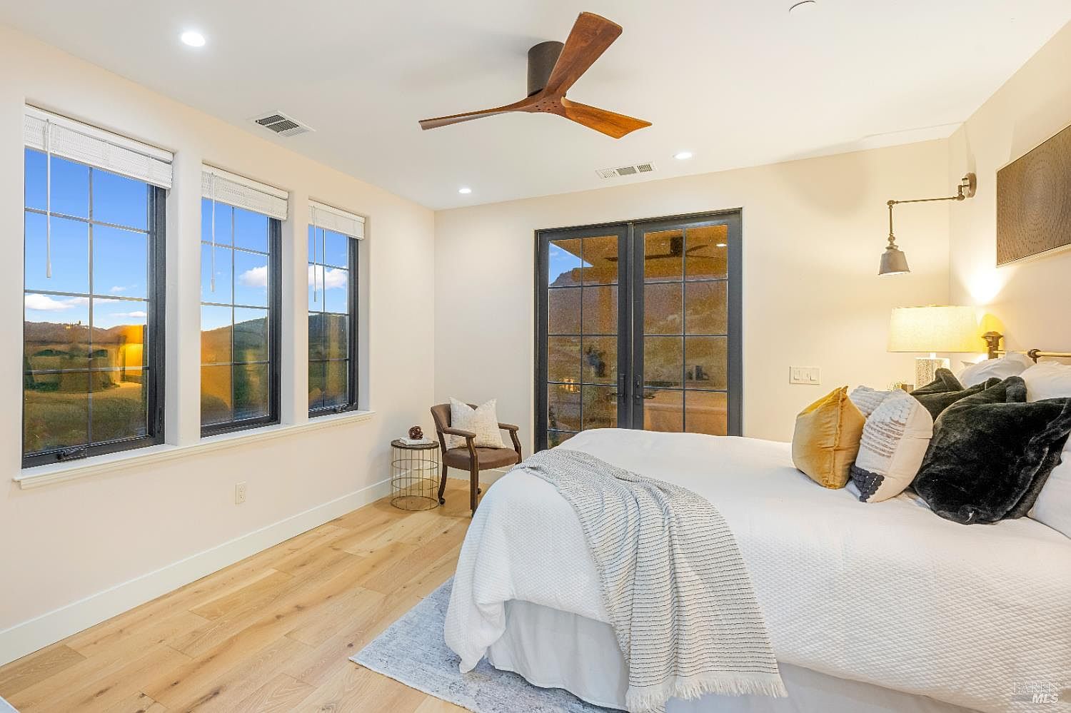 This is a well-lit primary bedroom featuring a king-size bed with white linens, decorative pillows, and a throw blanket. The room has three windows offering natural light and a view of the landscape, complemented by a modern ceiling fan and stylish wall sconce. A small accent chair and table add to the room's cozy and inviting atmosphere.