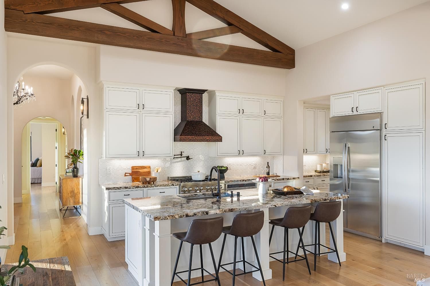 This is a bright and spacious kitchen featuring white cabinetry, a large island with seating, and stainless steel appliances. The kitchen has a high ceiling with wooden beams, adding a touch of rustic charm to the modern design. The perspective is from the side, showcasing the island and the surrounding cabinetry.
