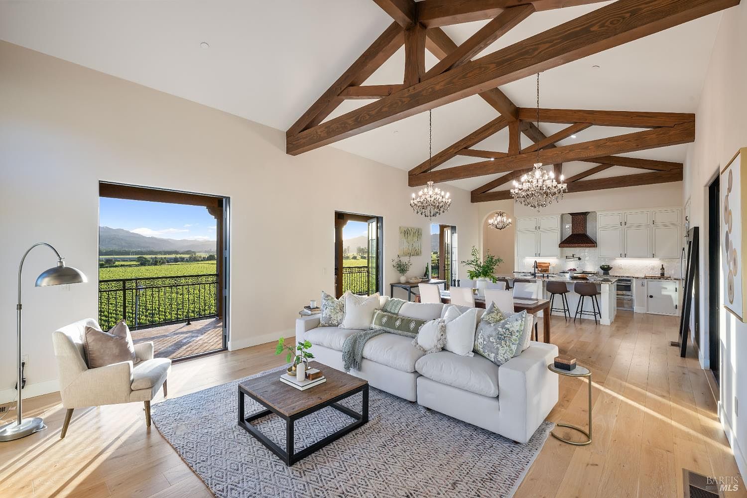 This is an interior shot of a luxurious living room that seamlessly blends into the dining and kitchen areas. The room features high vaulted ceilings with exposed wooden beams, a large white sectional sofa, and a stylish coffee table on a patterned rug. Large doors open to a balcony with views of a vineyard, creating an open and airy atmosphere.