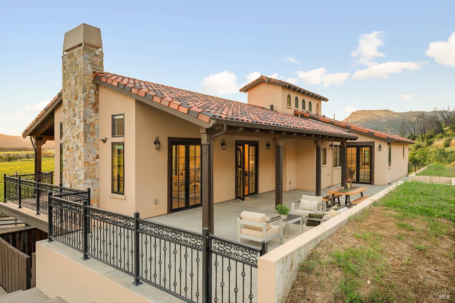 This image showcases the rear exterior of a Mediterranean-style home, featuring a terracotta tile roof, stucco walls, and dark-framed windows and doors. A covered patio with outdoor seating provides an inviting space for relaxation, while a stone chimney adds a touch of rustic charm. The landscape includes a well-maintained lawn and hillside vegetation.