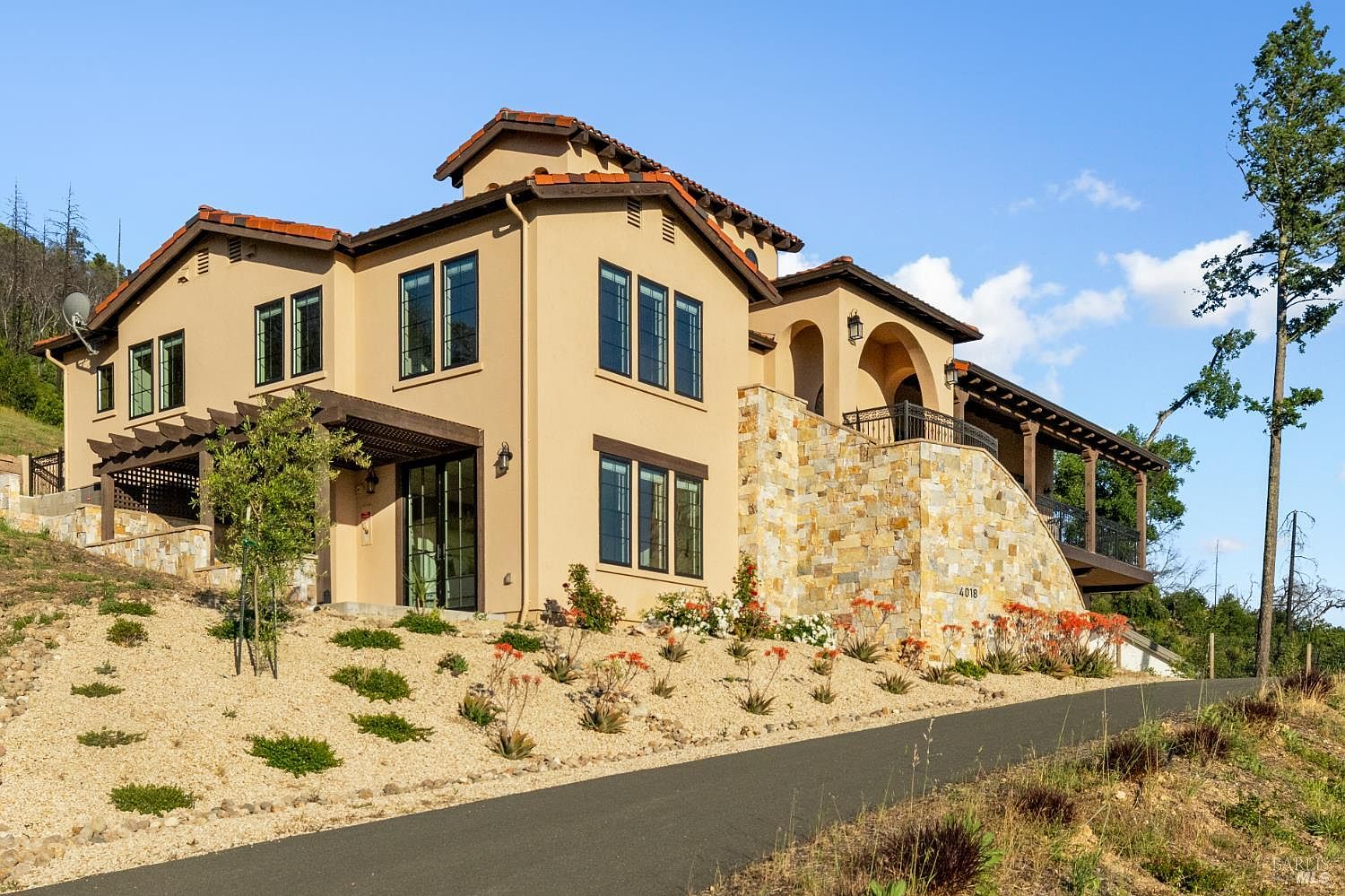 This is a front exterior view of a two-story Mediterranean-style home. The house features a stucco exterior, a red tile roof, and dark-framed windows. The landscaping includes gravel, shrubs, and flowers, and a paved driveway leads up to the house.
