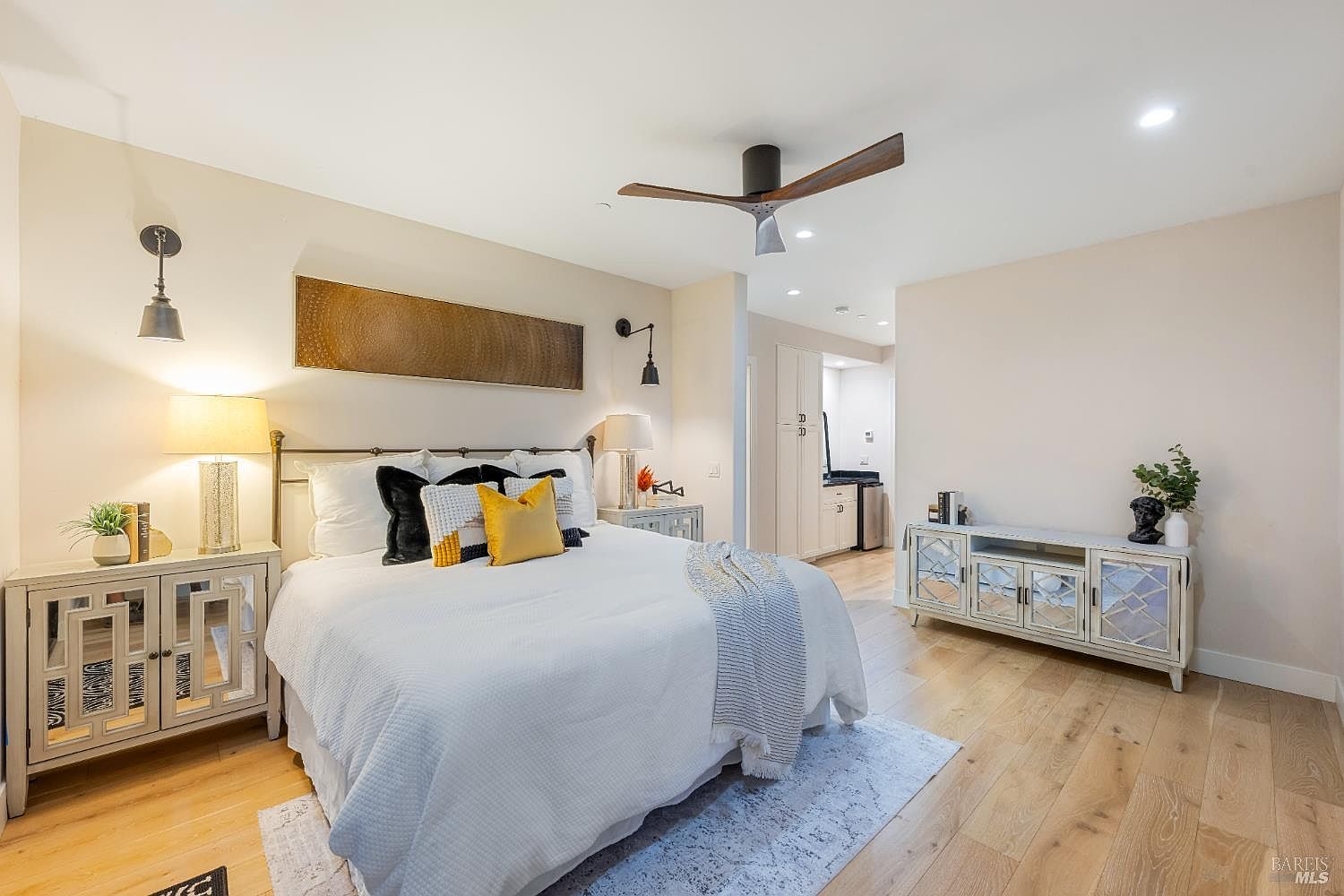 This is a well-lit primary bedroom featuring a queen-sized bed with white linens and decorative pillows. The room is furnished with mirrored nightstands and a matching console table, adding a touch of glamour. Hardwood floors and neutral wall colors create a warm and inviting atmosphere.