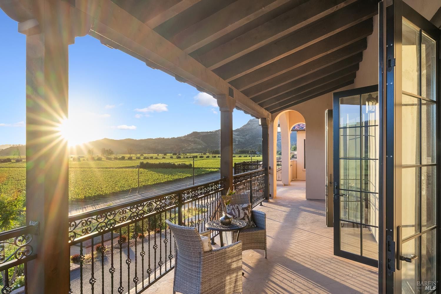 This image showcases a luxurious balcony overlooking a picturesque landscape of vineyards and distant mountains, bathed in the warm glow of the setting sun. The balcony features elegant wrought iron railings, comfortable wicker seating, and a covered area with exposed wooden beams, creating an inviting outdoor living space. The open doors lead to the interior, seamlessly blending indoor and outdoor living.