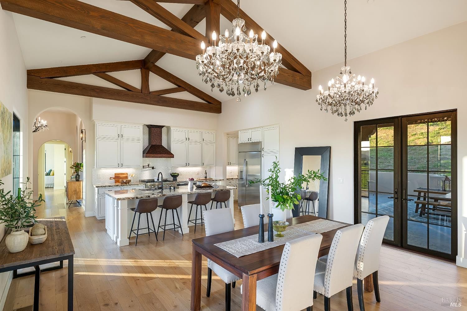 This is an interior shot of a dining room and kitchen area. The dining room features a wooden table with upholstered chairs and a decorative table runner, illuminated by a chandelier. The kitchen has white cabinets, granite countertops, and stainless steel appliances, with a large island and bar stools. The room has high ceilings with exposed wooden beams and a large window/door leading to the outside.