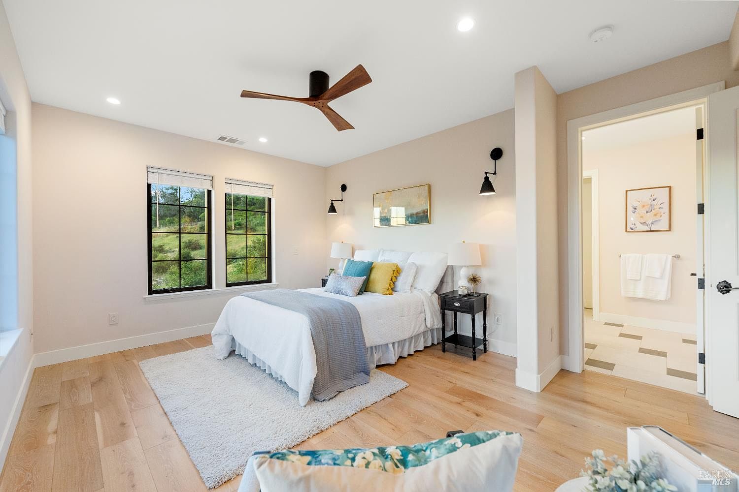 This is a bright and airy bedroom featuring hardwood floors, neutral-toned walls, and a ceiling fan. The room is furnished with a bed dressed in white linens and accented with colorful pillows and a gray throw. Natural light streams in through two windows, and the room opens to an en-suite bathroom, creating a comfortable and inviting space.