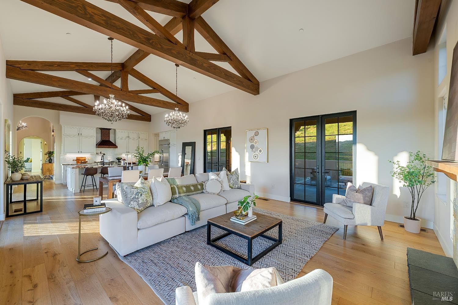 This is an interior shot of a spacious living room that seamlessly blends into an open-concept kitchen and dining area. The room features high vaulted ceilings with exposed wooden beams, elegant chandeliers, and large windows that provide ample natural light. The decor is a mix of modern and rustic, with a comfortable sectional sofa, a patterned area rug, and stylish accent furniture, creating an inviting and luxurious atmosphere.