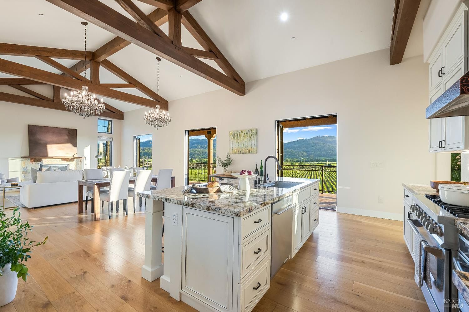 This is a bright and spacious kitchen featuring a large island with granite countertops, white cabinetry, and stainless steel appliances. The open floor plan seamlessly connects the kitchen to the dining and living areas, enhanced by exposed wooden beams and elegant chandeliers. Large windows and doors offer stunning views of the surrounding vineyards, creating a luxurious and inviting atmosphere.
