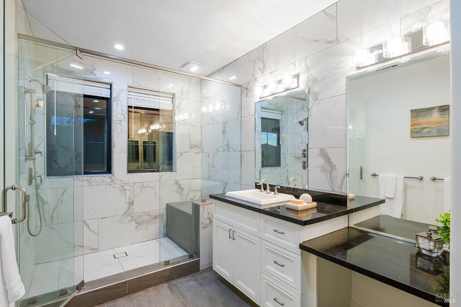 This is a well-lit primary bathroom featuring marble-like tile walls and a glass-enclosed shower. The vanity has a dark countertop and white cabinetry, complemented by a large mirror and modern lighting fixtures. The space feels clean, luxurious, and designed for comfort.