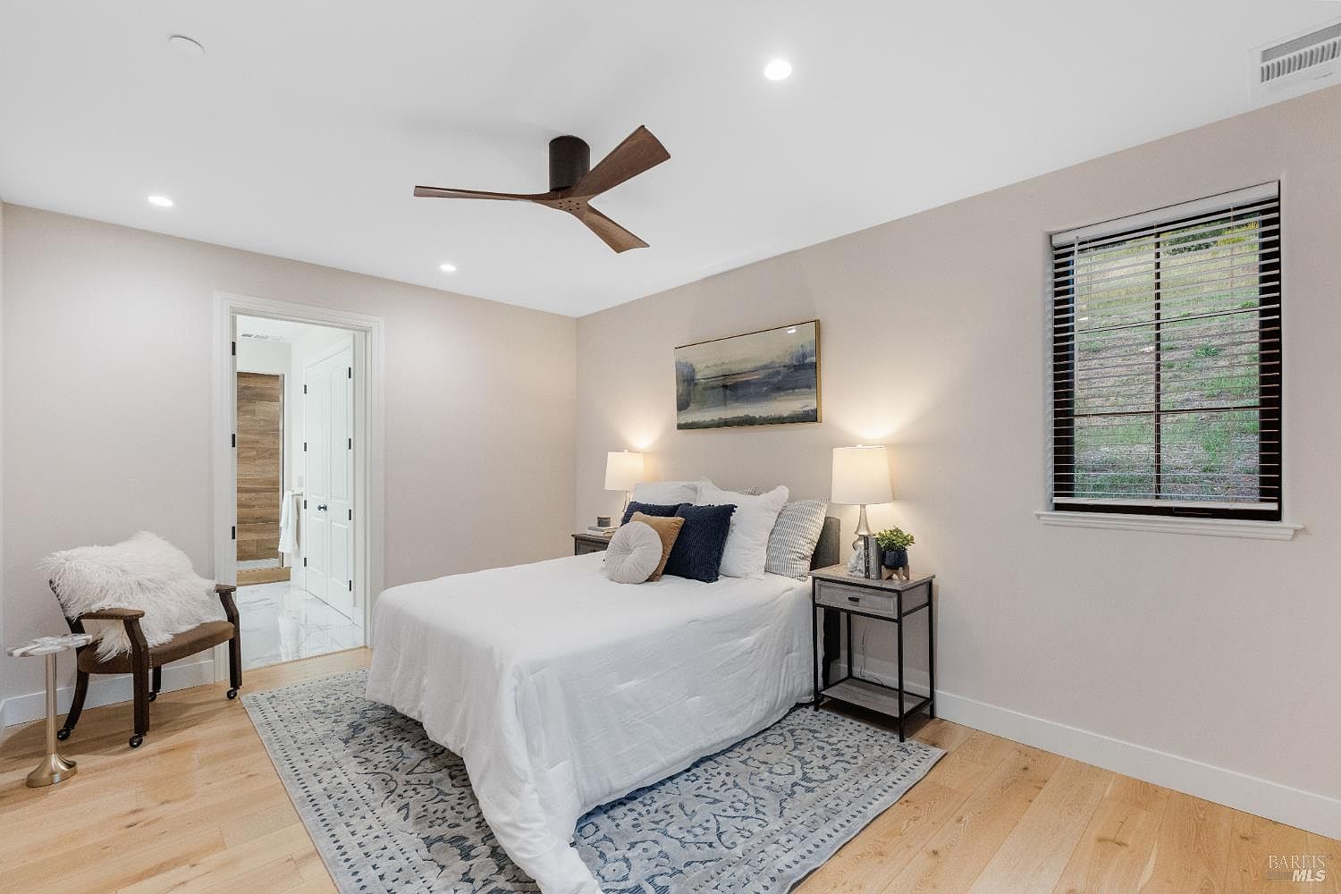 This is a well-lit bedroom featuring a bed with white linens, decorative pillows, and two side tables with lamps. An abstract painting hangs above the bed, and a window with blinds provides natural light. The room is decorated with a blue patterned rug and a chair with a fur throw, creating a cozy and inviting atmosphere.
