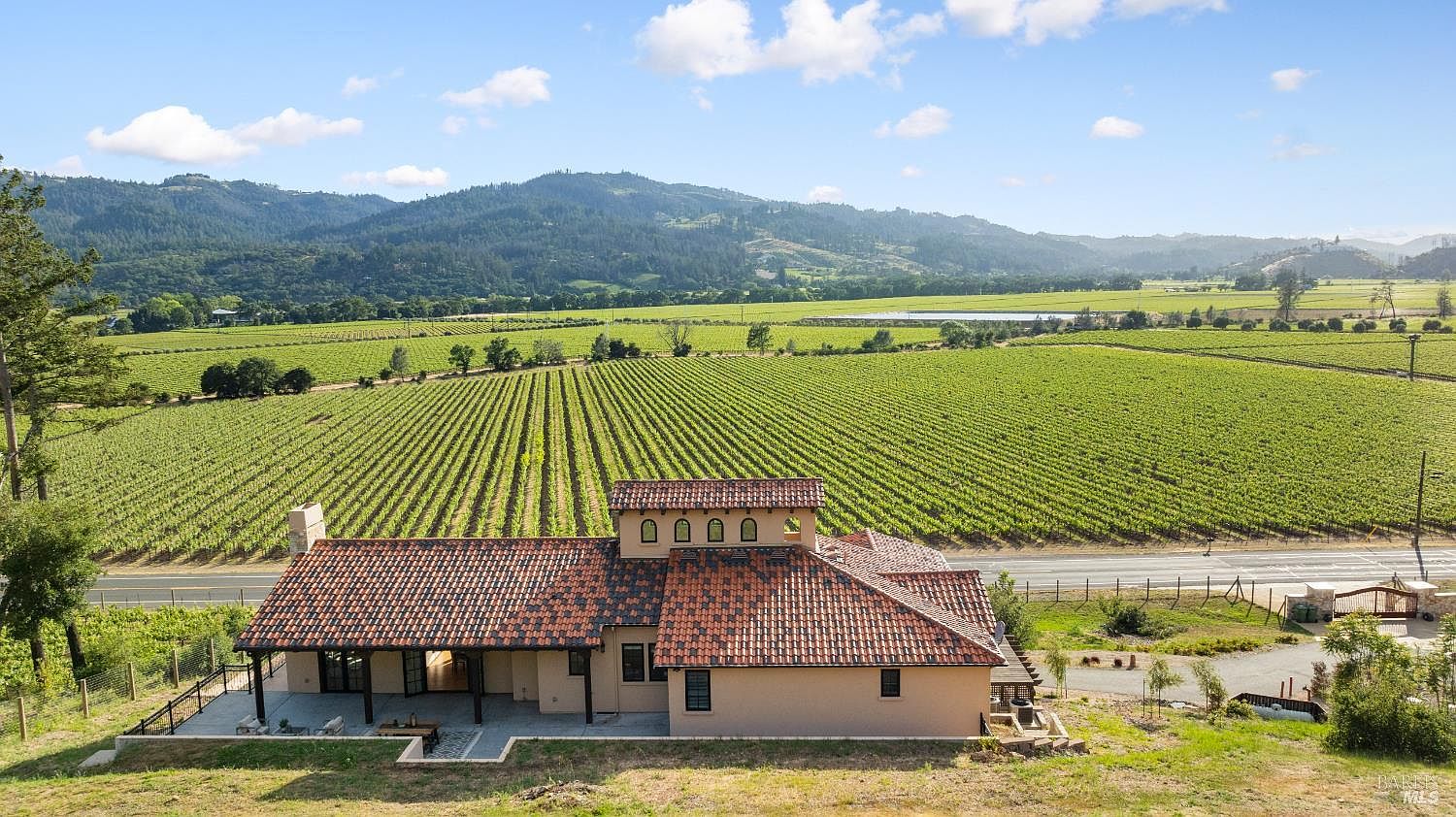 This aerial view showcases a stunning property nestled amidst a sprawling vineyard with rolling hills in the background. The building features a terracotta tile roof and a spacious patio area, blending seamlessly with the natural landscape. The overall impression is one of luxury and tranquility, ideal for a vineyard estate.