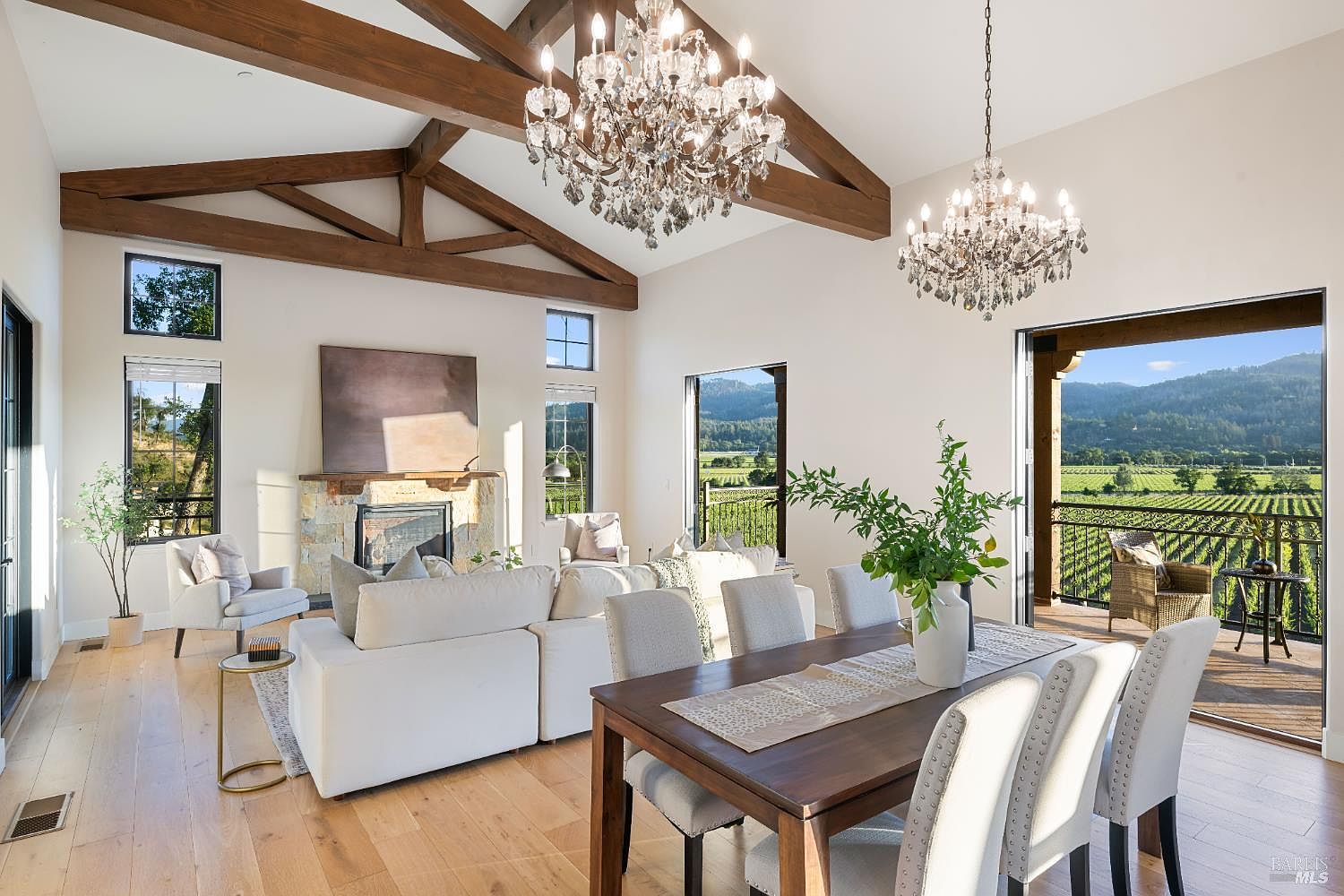 This is an interior shot of a luxurious living room and dining area featuring high vaulted ceilings with exposed wooden beams and two elegant crystal chandeliers. The room is bathed in natural light, showcasing a comfortable white sofa, a dining table set for six, and a stunning view of a vineyard through large sliding glass doors. The overall impression is one of spaciousness, elegance, and connection to the surrounding landscape.