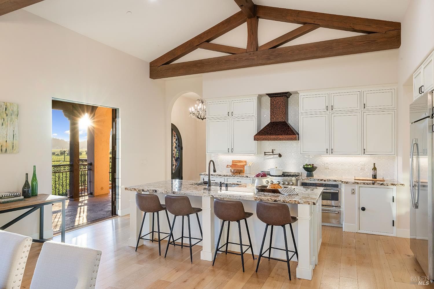 This is a bright and airy kitchen featuring a large island with seating, white cabinetry, and a decorative range hood. The kitchen has hardwood floors and exposed wooden beams on the ceiling, adding a touch of rustic charm. A large opening leads to an outdoor balcony, offering a view of the landscape and bringing natural light into the space.