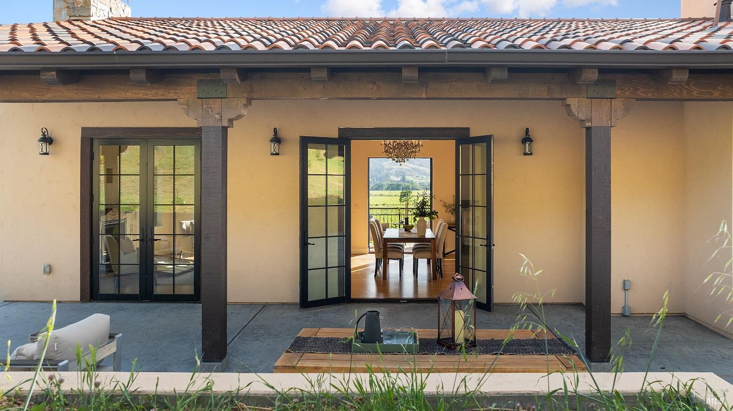 This image showcases a charming outdoor patio area, seamlessly blending indoor and outdoor living. The patio features a concrete floor, wooden beams, and a stylish outdoor seating arrangement with a wooden coffee table. Through the open French doors, a glimpse of the dining room with a chandelier and a scenic view of the landscape can be seen, creating an inviting and luxurious atmosphere.