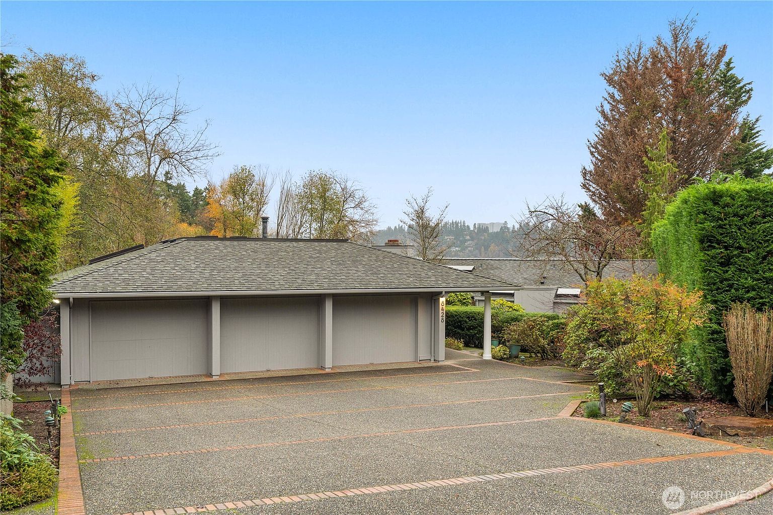 This image showcases a spacious three-car garage with a low-pitched, shingled roof and neutral-toned siding. A wide, textured concrete driveway leads up to the garage doors, framed by manicured hedges and mature trees that provide a sense of privacy. The perspective is a wide-angle shot from the driveway, capturing the functional layout and the surrounding lush landscaping.