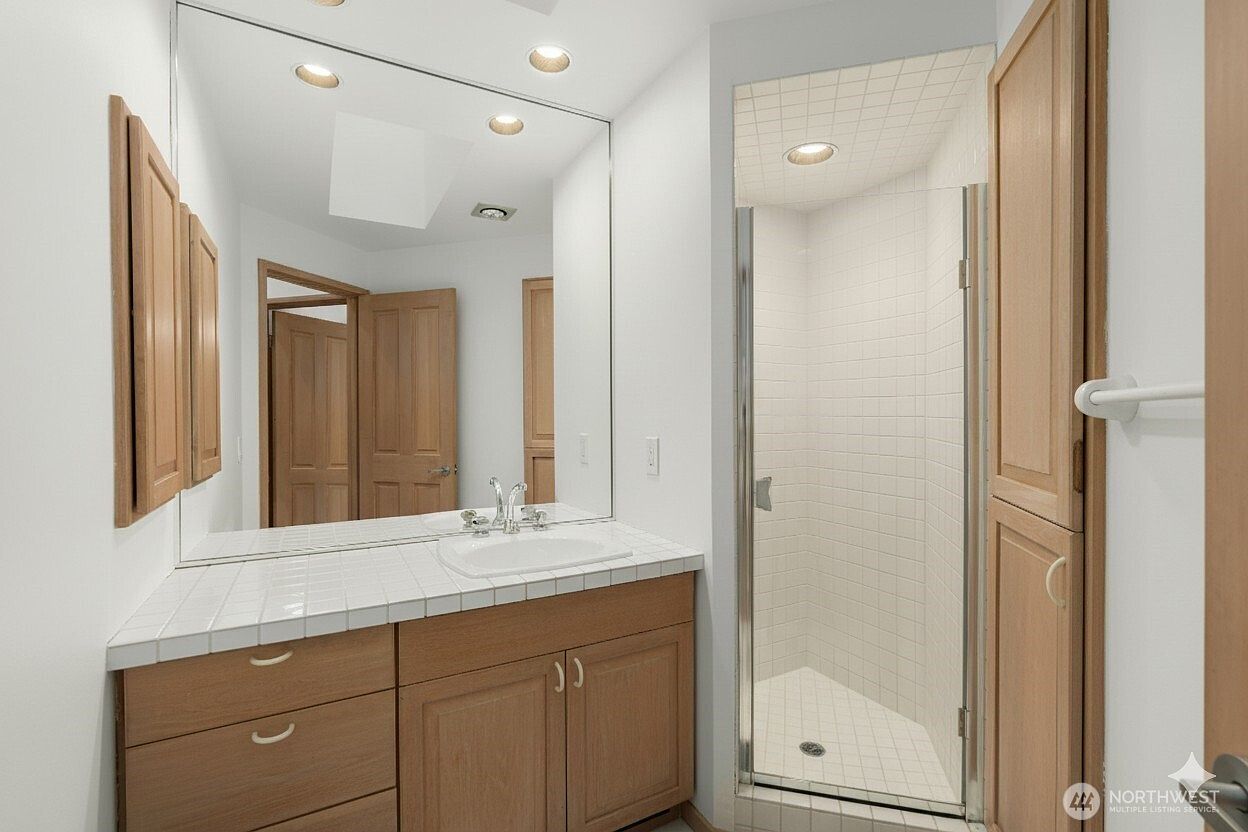 This bathroom features a light wood vanity with a white tiled countertop and a large, wall-mounted mirror. To the right, a glass-enclosed shower stall is finished with white wall tiles and a neutral-toned floor. The space is bright and functional, with a clean, traditional aesthetic.