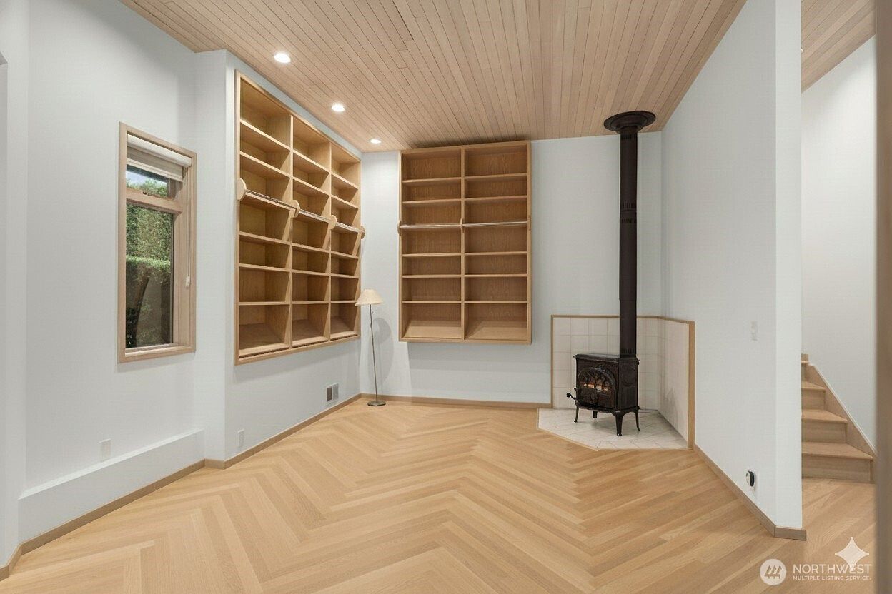 This bright study features light wood herringbone flooring and a matching wood-paneled ceiling, creating a warm and cohesive aesthetic. The room is equipped with extensive built-in shelving units and a freestanding cast-iron stove set on a tiled hearth in the corner. A tall window allows natural light to fill the space, which is framed by clean white walls and minimalist architectural lines.