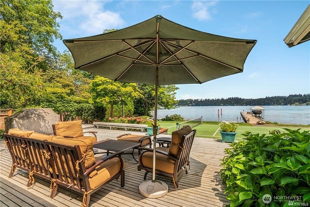 This expansive wooden deck offers a serene outdoor living space overlooking a calm lake. The area is furnished with comfortable patio seating under a large, dark green umbrella, providing a perfect spot for relaxation. Beyond the deck, a private tennis court and a wooden dock extending into the water enhance the property's recreational appeal.