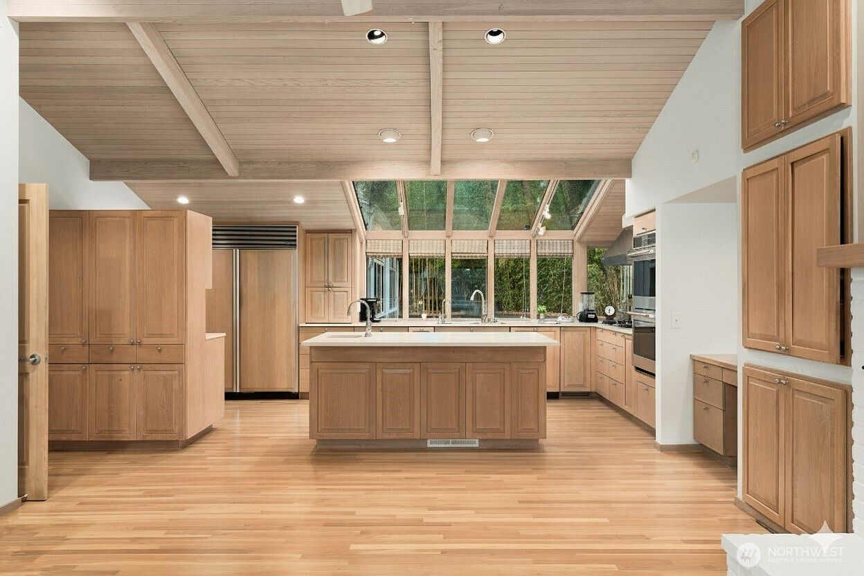 This spacious, light-filled kitchen features a central island with a light-toned countertop and extensive light wood cabinetry throughout. A dramatic sloped ceiling with exposed beams adds architectural interest, while a large wall of windows and a skylight-style glass roof section bring in abundant natural light. The open-concept layout creates an airy, inviting atmosphere, complemented by warm wood flooring and integrated stainless steel appliances.