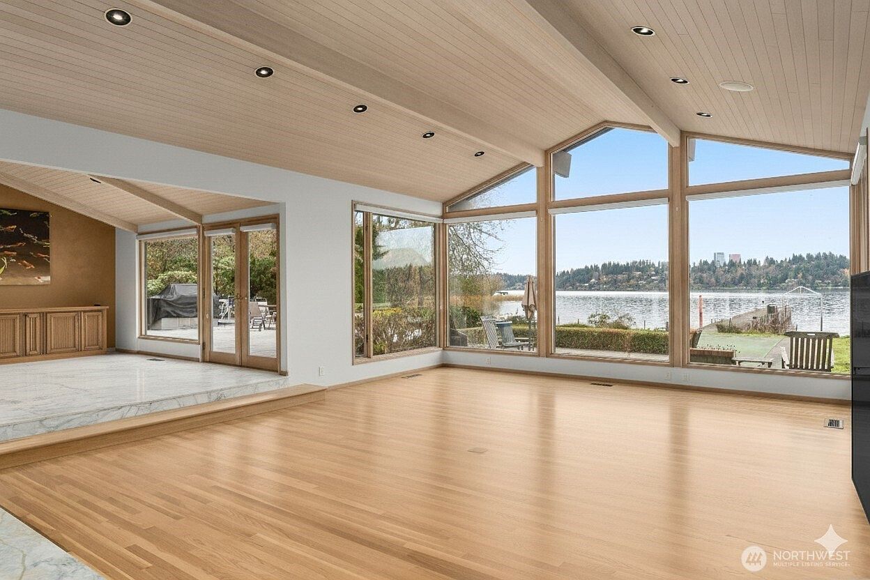 This spacious living area features light wood flooring and a vaulted, wood-paneled ceiling with recessed lighting. A wall of large windows provides an expansive, unobstructed view of the lake and surrounding landscape, while a raised platform area with light-colored stone flooring adds architectural interest. The room feels bright and airy, emphasizing a seamless connection between the interior living space and the natural scenery outside.