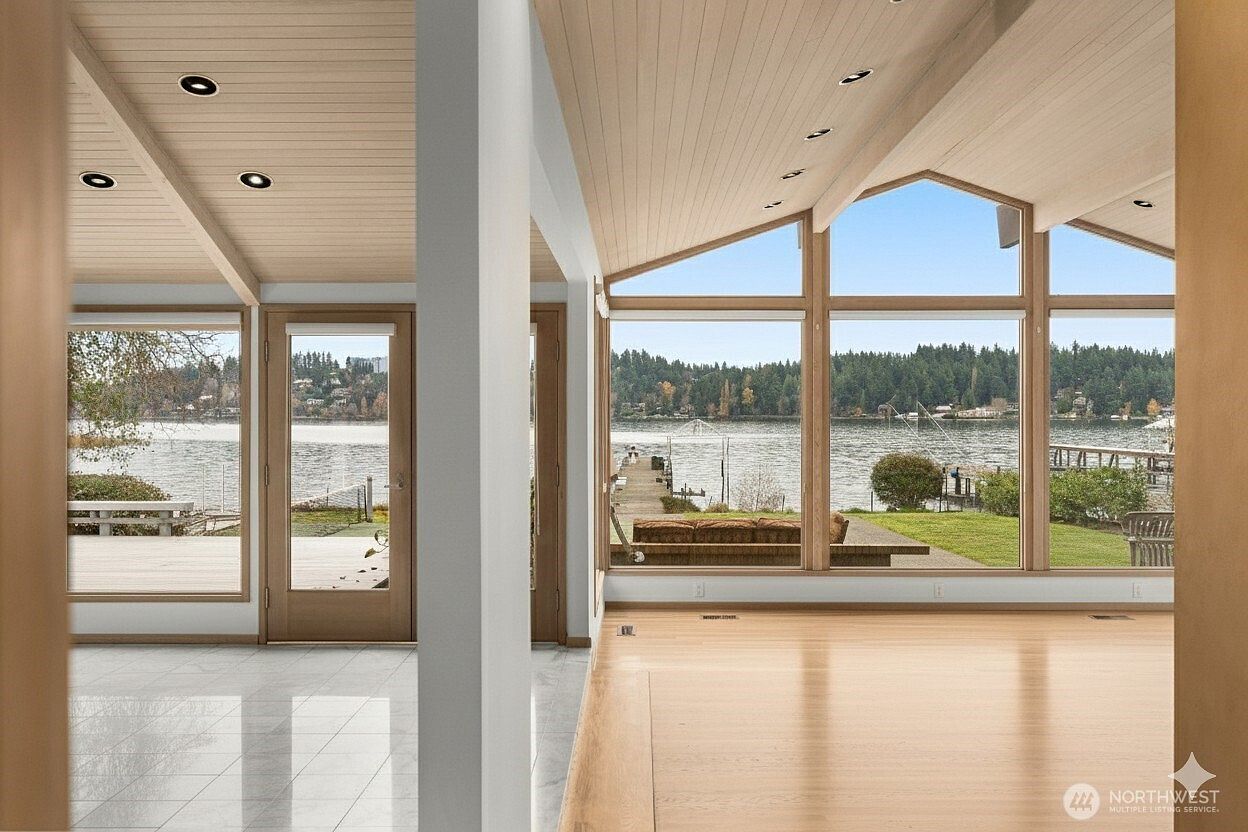 This bright living area features light wood flooring and a vaulted ceiling with recessed lighting, creating an airy and open atmosphere. Large windows provide an expansive, unobstructed view of the lake, a private dock, and the surrounding greenery. The perspective is from inside the home, looking out through the glass walls toward the water, emphasizing the seamless connection between the indoor living space and the natural landscape.