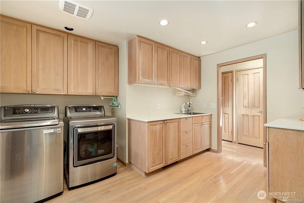 This functional laundry room features light wood cabinetry providing ample storage space above and below the counter. The room is equipped with stainless steel laundry appliances and a built-in sink, set against a neutral wall color and light-toned wood flooring. The perspective captures the entire workspace, highlighting the clean, organized layout and the transition into an adjacent room through a doorway.