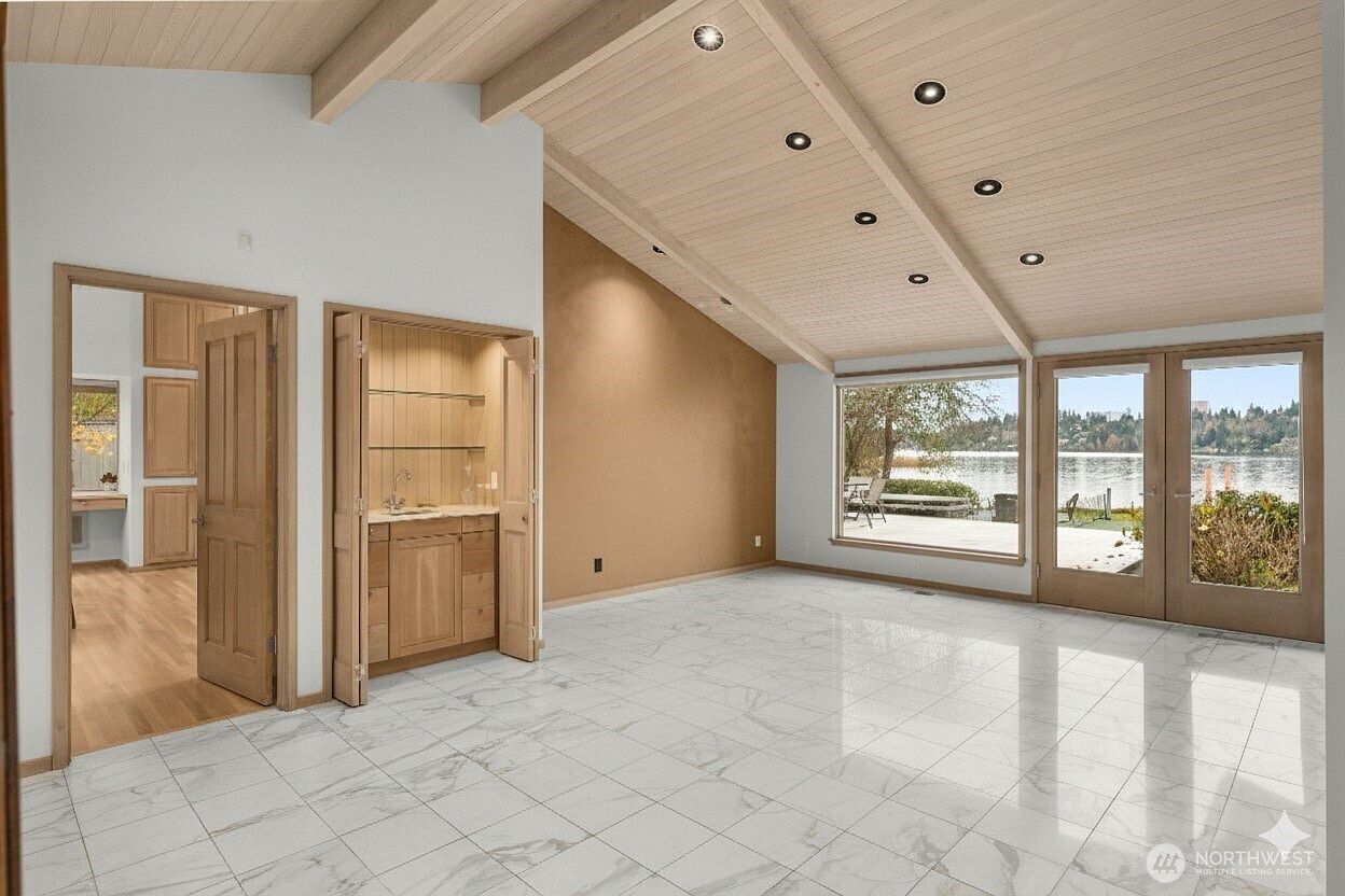 This bright living area features a vaulted ceiling with light wood paneling and recessed lighting, creating an airy and spacious atmosphere. The room is finished with light-colored patterned tile flooring and includes a built-in wet bar tucked into a recessed alcove. Large windows and glass doors provide an expansive view of the water, seamlessly connecting the interior space to the outdoors.