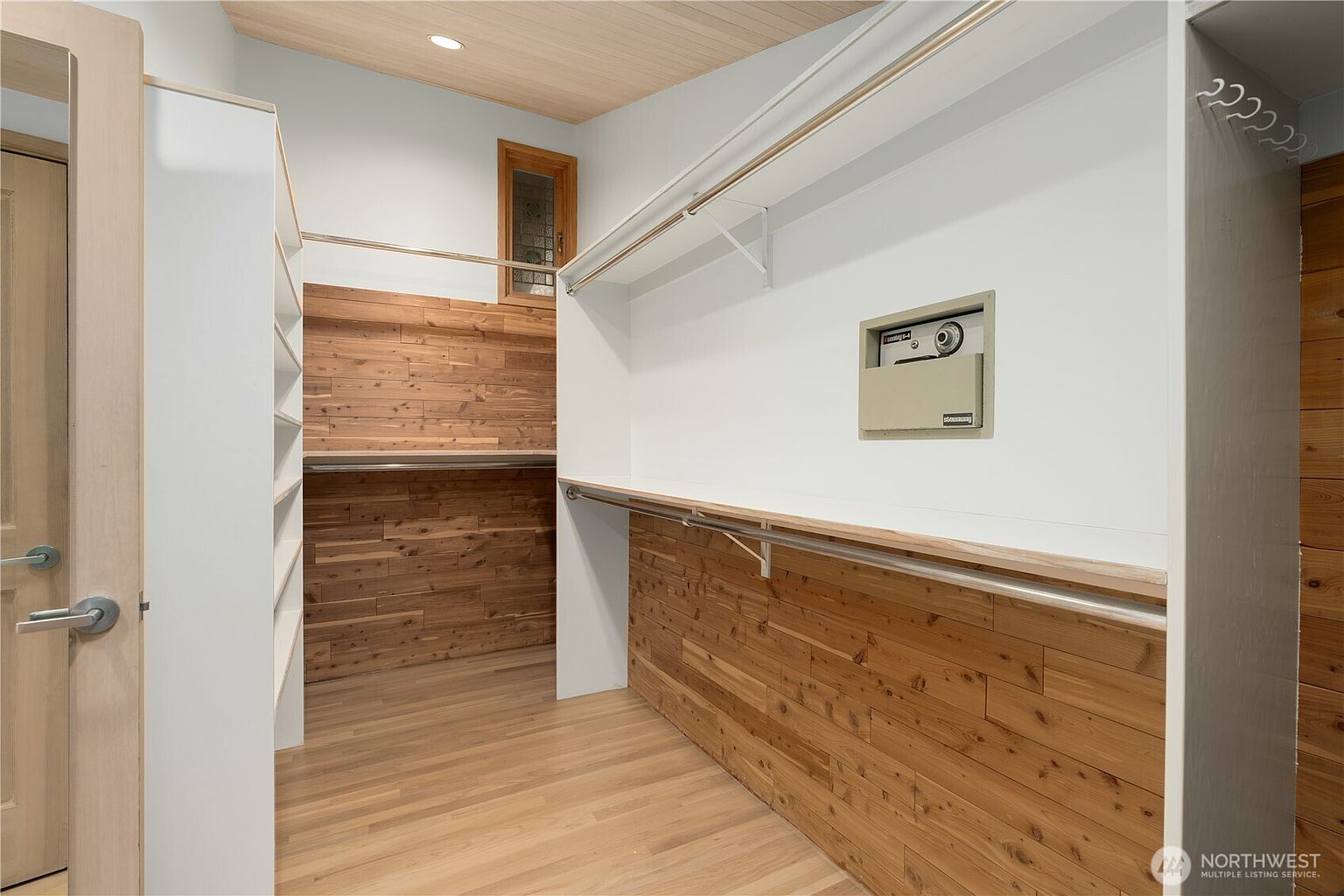 This walk-in closet features a blend of light wood flooring and horizontal wood-plank wall accents, creating a warm and textured aesthetic. The space is equipped with white shelving units, a hanging rod, and a built-in wall safe, offering organized storage solutions. A small, high-placed window adds natural light, while the clean lines and neutral color palette provide a modern and functional feel.