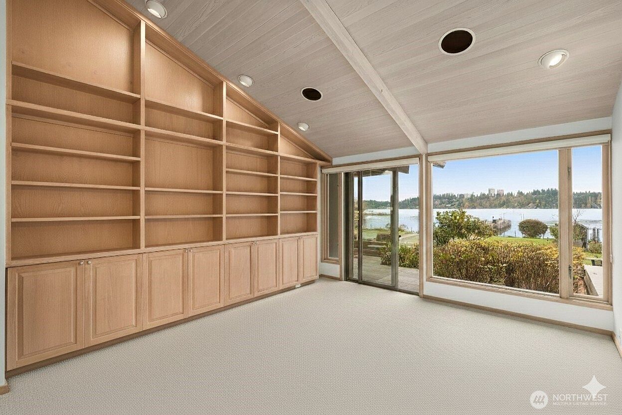 This bright room features a large, built-in light wood shelving unit that spans the length of one wall, providing ample storage and display space. A sliding glass door and a large window offer expansive views of the lake and surrounding greenery, filling the space with natural light. The room is finished with neutral carpeting and a vaulted ceiling, creating an airy and versatile atmosphere suitable for a home office or library.