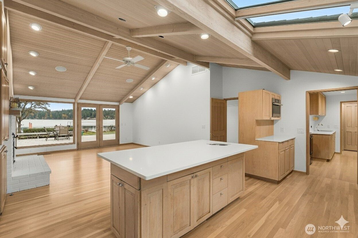 This open-concept kitchen and living area features a large central island with a light-colored countertop and light wood cabinetry. The space is defined by a vaulted wood-paneled ceiling with exposed beams, a skylight, and large glass doors that provide a view of the water outside. The room has a bright, airy feel with light wood flooring and recessed lighting throughout.