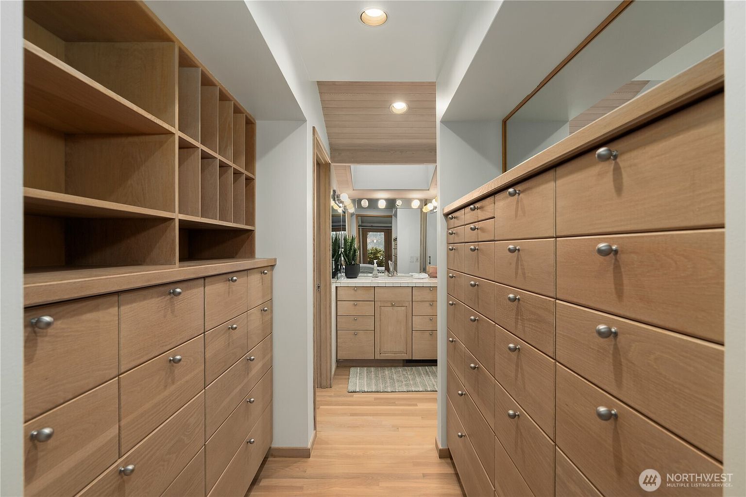 This walk-in closet features extensive custom light wood cabinetry with numerous drawers and open shelving, providing ample storage space. The design leads directly into a bathroom, creating a seamless transition between the dressing area and the vanity. The space is illuminated by recessed lighting and maintains a clean, organized, and modern aesthetic.